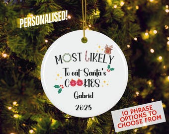 May include: A round white ceramic Christmas ornament featuring the text "MOST LIKELY To eat Santa's COOKIES" in black, with a small reindeer, candy cane, holly, and red ornaments. The name "Gabriel" and the year "2025" are printed below.