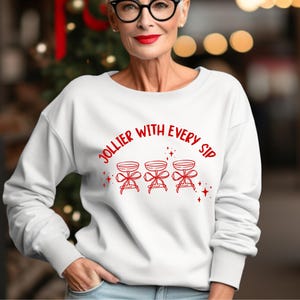 May include: A white long-sleeve crewneck sweatshirt featuring a red graphic print of three champagne coupes tied with bows and the text "JOLLIER WITH EVERY SIP" arched above the glasses, accented with small red starbursts and sparkles.