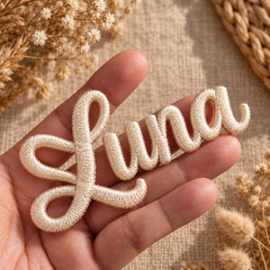 Op de afbeelding: Een crèmekleurige geborduurde patch met de naam Luna in een cursief, afgerond lettertype. De letters hebben een verhoogd, getextureerd uiterlijk met dicht stiksel, wat een zacht, driedimensionaal effect op het oppervlak van de stof creëert.