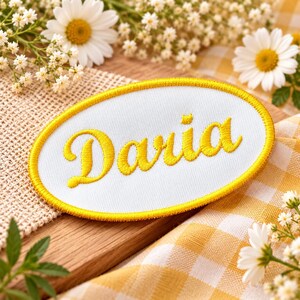 May include: An oval-shaped embroidered patch featuring a white fabric base with a bright yellow stitched border. The name Daria is embroidered in the center in a bold, cursive yellow script font with a distinct yellow thread texture.