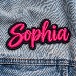 Peut inclure: Un écusson brodé rose vif affichant le prénom Sophia en écriture cursive avec une bordure noire épaisse, fixé sur un tissu en jean bleu clair avec des coutures visibles et une couture horizontale.