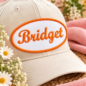 May include: A beige baseball cap featuring an oval patch on the front. The patch has a white background with a bright orange embroidered border and the name Bridget written in matching orange cursive script across the center.