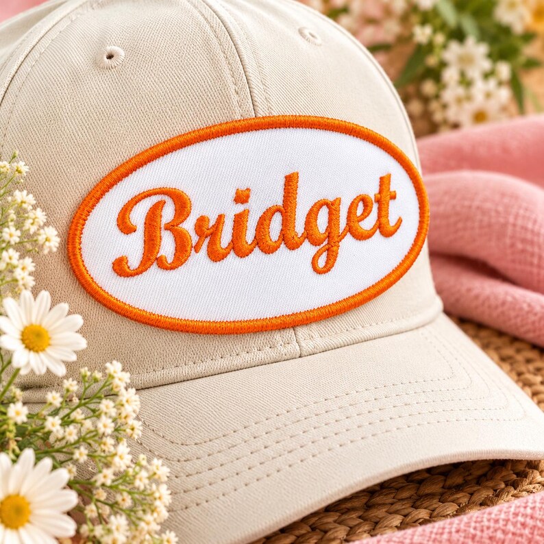 May include: A beige baseball cap featuring an oval patch on the front. The patch has a white background with a bright orange embroidered border and the name Bridget written in matching orange cursive script across the center.