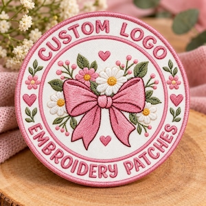 May include: A circular embroidered patch with a white background and a pink border. The text CUSTOM LOGO EMBROIDERY PATCHES is stitched in pink. The center features a large pink bow, white daisies, pink flowers, green leaves, and small pink hearts.