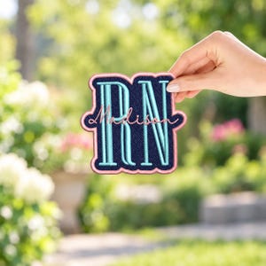 May include: A navy blue embroidered patch featuring the light blue letters RN with the name Madison stitched in pink cursive script across the center, all outlined with a pink border in a scalloped shape.