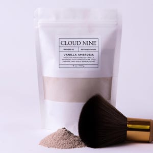 May include: A white pouch of "Cloud Nine" setting powder, with the text "Vanilla Ambrosia." A pile of powder and a makeup brush with a wooden handle and gold ferrule are next to the pouch. The brush has dark brown bristles.