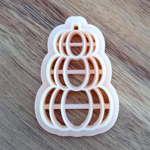 May include: A light pink and orange pumpkin-shaped cookie cutter. The cutter is made of plastic and has a stacked pumpkin design. The cutter is on a wooden surface.