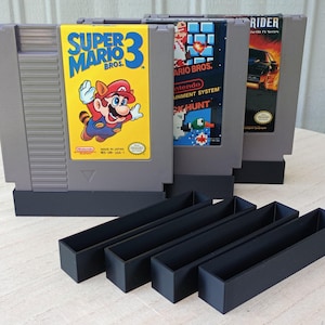 May include: Three gray Nintendo Entertainment System game cartridges standing upright in black plastic display stands. The cartridges feature labels for Super Mario Bros. 3, Super Mario Bros./Duck Hunt, and Knight Rider. Four empty black stands are in front.