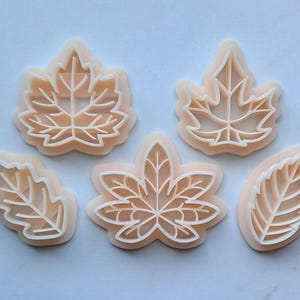 May include: Five plastic leaf-shaped cookie cutters. The cutters are in various shapes and sizes, including maple, oak, and other leaf designs. The cutters are a light peach color.