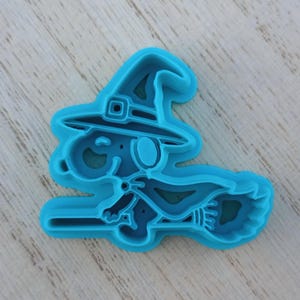 May include: A bright blue plastic cookie cutter shaped like a cartoon dog wearing a pointed witch hat and a cape while riding a broomstick. The design features raised edges for cutting dough and a detailed outline of the character's face and body.