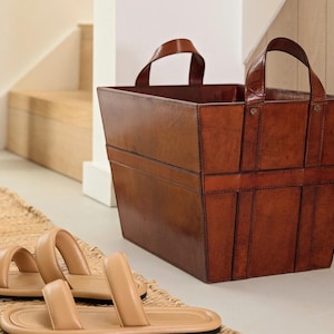 May include: A brown leather storage basket with two handles. The basket is sitting on a white floor with a tan rug in the foreground.