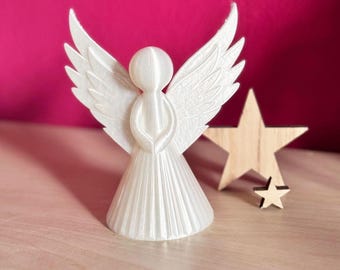 Pode incluir: Uma estatueta de anjo branca impressa em 3D com asas abertas, ao lado de uma estrela de madeira. O anjo tem uma túnica fluida e asas detalhadas. O fundo é rosa vibrante.