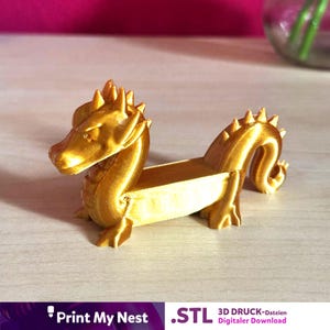 May include: A gold-coloured dragon ornament with a textured finish. The dragon features a detailed head, a long body, and a small trough. The figurine is likely 3D-printed and is a decorative item.