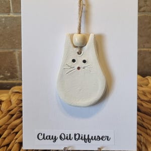 May include: A white clay cat-shaped oil diffuser with black eyes and a red nose, suspended by a natural twine cord and a wooden bead. The text "Clay Oil Diffuser" is printed on a white card. The diffuser is a decorative item.