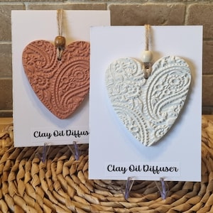 May include: Two heart-shaped clay oil diffusers. One is terracotta, the other is white. Both feature a paisley design and are on white cards with the text "Clay Oil Diffuser". They are suspended by twine.