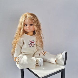 May include: A doll with long blonde hair wearing a cream-colored sweatshirt and ripped pants. The sweatshirt has a graphic of red sneakers and the text "STYLE AUTHENTIC WEAR". The doll is wearing white sneakers with black soles.