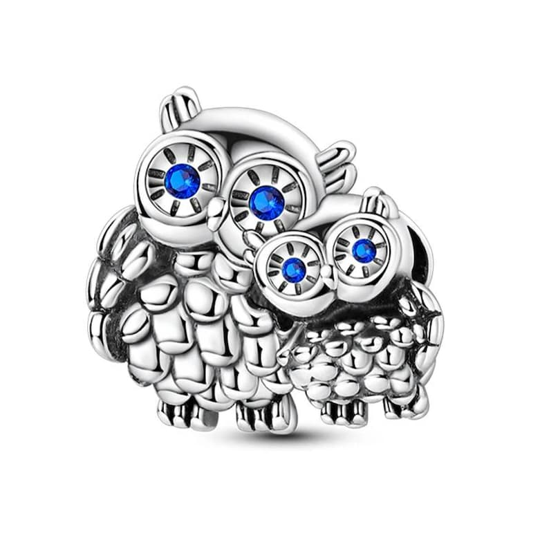 K&ouml;nnte beinhalten: Ein silberner Charm-Anh&auml;nger mit zwei Eulen. Die Eulen haben gro&szlig;e, runde Augen mit blauen Edelsteinzentren. Der Charm hat eine detaillierte, strukturierte Oberfl&auml;che und ist wahrscheinlich ein Anh&auml;nger oder eine Perle f&uuml;r ein Armband.