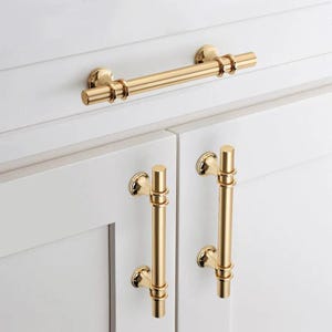 May include: Gold-toned cabinet hardware, including a long bar handle and two vertical pull handles, are mounted on white cabinet doors. The hardware has a cylindrical design with decorative ring accents, offering a classic and elegant aesthetic.