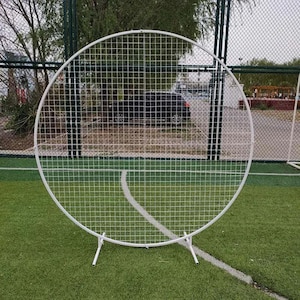 Puede incluir: Un telón de fondo de rejilla circular de metal blanco para eventos. La rejilla está soportada por un soporte con tres patas. El telón de fondo está colocado sobre un campo verde con una línea blanca.