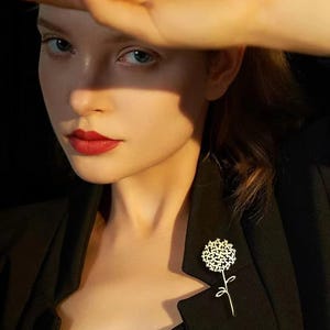 Peut inclure: Broche dorée en forme de fleur, épinglée sur un blazer noir. La broche présente un motif floral détaillé avec une tige et des pétales délicats. L'image est un gros plan, soulignant les détails complexes de la broche et le contraste avec le tissu sombre.