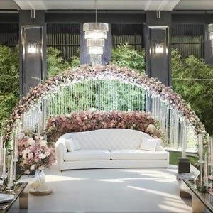 Può includere: Allestimento per una cerimonia nuziale con un divano bianco trapuntato sotto un arco floreale. L'arco è adornato con fiori rosa e bianchi e fili di cristallo sospesi. Un lampadario è appeso sopra e lo sfondo comprende vegetazione e illuminazione decorativa.