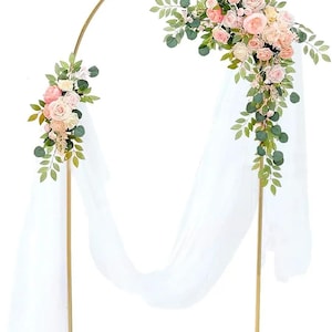 Könnte beinhalten: Zwei goldfarbene Hochzeitsbögen aus Metall, jeweils mit halbkreisförmiger Oberseite und rechteckiger Basis. Die Bögen sind mit rosa und weißen Blumen, grünen Blättern und weißem Stoff dekoriert.