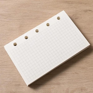 Puede incluir: Una pila de papel cuadriculado blanco con un patrón de cuadrícula y cuatro agujeros perforados a lo largo del borde superior. El papel está sobre una superficie de madera de color marrón claro.