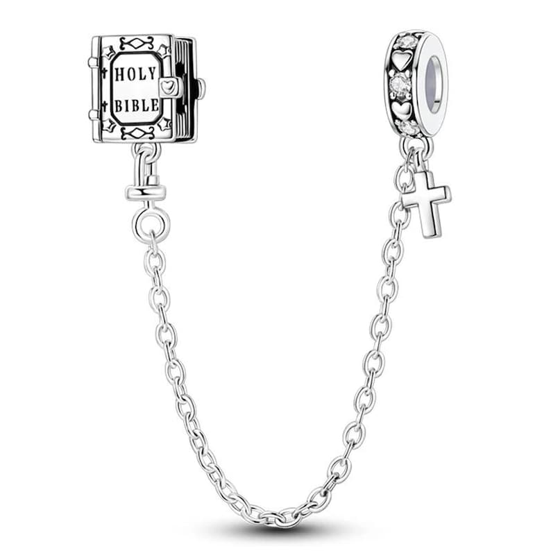 K&ouml;nnte beinhalten: Silberne Charm-Armband-Sicherheitskette mit einem Holy Bible-Charm und einem Charm mit Herzen und klaren Steinen. Ein kleiner Kreuz-Charm h&auml;ngt an dem Charm mit Herzen. Die Kette verbindet die Charms.