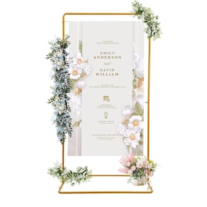 Könnte beinhalten: Ein goldfarbenes Hochzeitszeichen aus Metall mit Blumendekorationen. Auf dem Schild stehen die Namen "Emily Anderson und David William" und Hochzeitsdetails. Weiße und rosa Blumen und Grün schmücken das Schild.