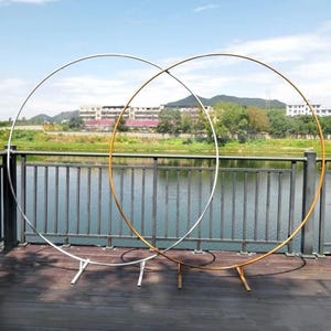 Può includere: Due grandi archi nuziali circolari in metallo. Uno è argentato, l'altro dorato. Ogni arco è sostenuto da una base con gambe. Gli archi sono allestiti all'aperto vicino a uno specchio d'acqua e a una terrazza in legno.