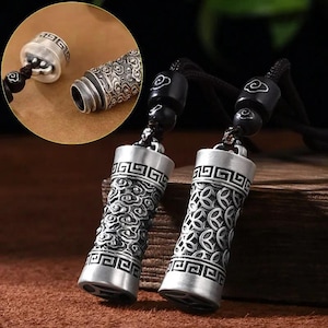 Op de afbeelding: Twee zilverkleurige cilindrische hangers met ingewikkelde wolken- en geometrische patronen. Elke hanger is bevestigd aan een zwart koord met zwarte kralen. Eén hanger is losgemaakt en toont de binnenkant. Waarschijnlijk voor sieraden of decoratieve doeleinden.