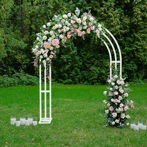 May include: A white metal wedding arch decorated with pink and white flowers. The arch is set on a green lawn with candles in glass holders. The arch is a decorative element for outdoor ceremonies.