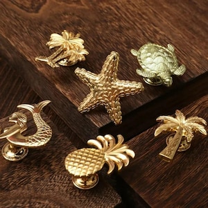 May include: Six gold-toned metal cabinet knobs with a beach theme. The knobs feature a pineapple, palm tree, mermaid, starfish, turtle, and another palm tree.