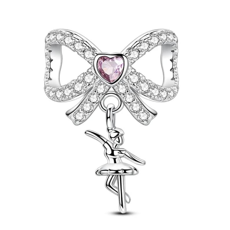K&ouml;nnte beinhalten: Silberner Charm mit einem Bogen-Design, besetzt mit klaren Kristallen und einem rosa herzf&ouml;rmigen Edelstein. Ein Ballerina-Charm h&auml;ngt darunter.
