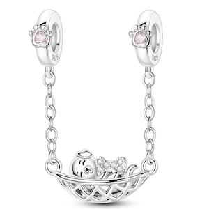 K&ouml;nnte beinhalten: Eine silberne Charm-Armband-Sicherheitskette mit einem Design eines schlafenden Tieres in einem Korb. Der Charm-Anh&auml;nger zeigt ein kleines Tier mit einer Schleife und einem Korb. Die Kette hat zwei Pfotenabdruck-Charms mit rosa herzf&ouml;rmigen Edelsteinen.