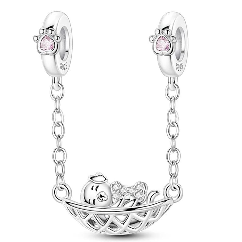 K&ouml;nnte beinhalten: Eine silberne Charm-Armband-Sicherheitskette mit einem Design eines schlafenden Tieres in einem Korb. Der Charm-Anh&auml;nger zeigt ein kleines Tier mit einer Schleife und einem Korb. Die Kette hat zwei Pfotenabdruck-Charms mit rosa herzf&ouml;rmigen Edelsteinen.