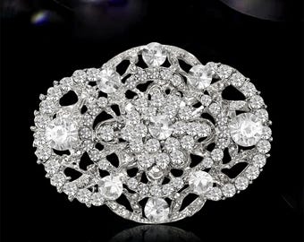 May include: Silver-toned oval brooch with a detailed floral design. The brooch is encrusted with numerous clear, sparkling crystals of varying sizes, creating a dazzling effect. The intricate openwork design adds to its elegance, set against a dark background.