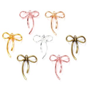 Puede incluir: Colección de siete colgantes de lazo metálicos en varios colores, incluyendo dorado, oro rosa, plateado y bronce. Los colgantes están dispuestos sobre un fondo blanco. Cada lazo tiene un bucle en la parte superior para sujetarlo a joyas u otras manualidades.