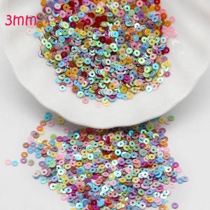 Puede incluir: Una colección de lentejuelas redondas de 3 mm en varios tonos pastel, incluyendo rosa, azul, verde y amarillo. Las lentejuelas están dispersas sobre un plato y una superficie blancos, ideales para manualidades y adornos.