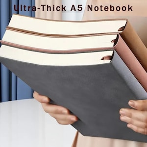 Op de afbeelding: Drie A5-notitieboeken met dikke pagina's worden vastgehouden. Het bovenste notitieboek is grijs, de middelste roze en de onderste bruin. De tekst "Ultra-Thick A5 Notebook" staat bovenaan de afbeelding.