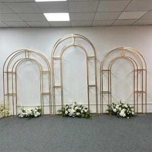 May include: Three gold metal arch wedding backdrops with a rectangular base and a rounded top. Each arch has a different design. White flowers and greenery are placed at the base of each arch, creating a decorative display.