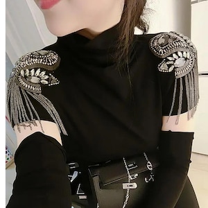 May include: Black turtleneck top with embellished shoulder pads featuring silver-tone chains and crystal accents. The top has long sleeves and is paired with a black purse with a silver chain.