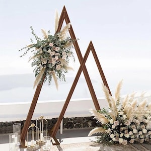 May include: A wooden wedding arch in a triangular design, decorated with floral arrangements. The arch is made of dark brown wood and decorated with white and pink roses, pampas grass, and greenery. The scene includes gold candle holders and a clear sky.