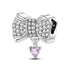 K&ouml;nnte beinhalten: Ein silberner Charm-Anh&auml;nger mit Schleifendesign, besetzt mit klaren Kristallen. Ein kleiner rosa herzf&ouml;rmiger Edelstein h&auml;ngt an der Schleife. Der Charm ist f&uuml;r ein Armband oder eine Halskette konzipiert.