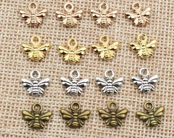 May include: A collection of small bee-shaped charms in various metallic finishes, including gold, silver, and bronze. Each charm features detailed wings and a loop for attaching to jewelry or crafts. The charms are arranged on a textured, tan fabric.