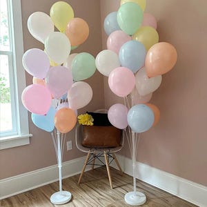 Puede incluir: Un conjunto de globos de colores pastel, incluyendo rosa, azul, amarillo y melocotón, dispuestos sobre soportes blancos. Los globos están agrupados, creando un ambiente festivo. Una silla y una ventana se ven al fondo.