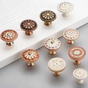 May include: A collection of decorative cabinet knobs in various colors, including bronze, gold, and white. Each knob features a floral or circular design, with a detailed, raised pattern. The knobs are mounted on a light-colored surface, showcasing their intricate details.