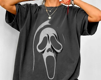 Może przedstawiać: Ciemnoszara koszulka z białą grafiką maski Scream. Oversize'owy t-shirt ma krótkie rękawy i okrągły dekolt. Grafika to stylizowana, otwartoustna twarz z dużymi oczami. Koszulka jest w stylu casual i wygodnym.