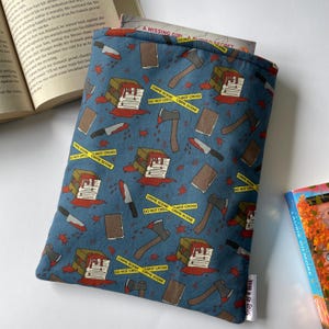 May include: A blue book sleeve featuring a pattern of axes, knives, books, and crime scene tape. The design includes red blood splatters and the words "Crime Scene" and "Do Not Cross".