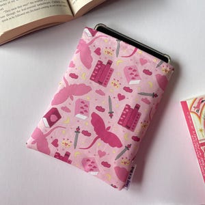 May include: Pink e-reader sleeve featuring a pattern of dragons, books, swords, crowns, and hearts. The sleeve is designed to hold an e-reader. The text "Snug as a Book" is visible. The sleeve is made of fabric.
